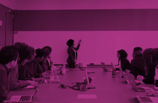 As a banner for the page, this is a photo with a purple filter overlay. The photo shows a group of people of different ethnicities sitting in a conference room. They're all looking at the front, where a woman with an afro, wearing a business suit, is explaining some concepts on a whiteboard.